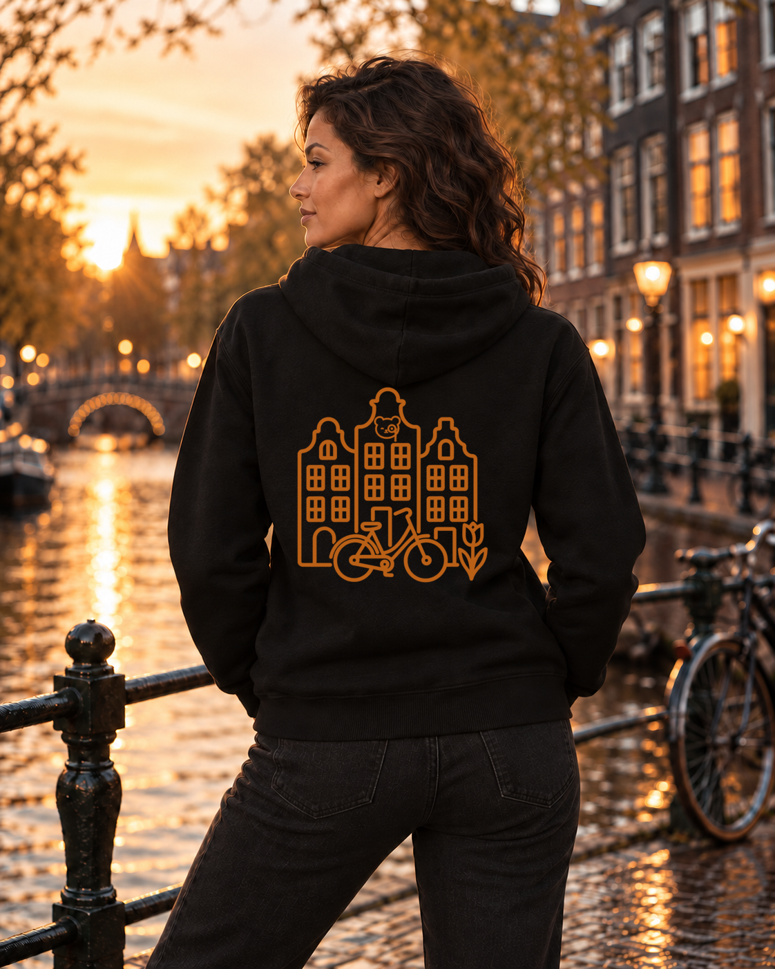 A woman stands on a canal-side street at sunset wearing a black hoodie with an orange graphic of canal houses and bicycles on the back, paired with dark jeans.